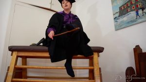 Pandora sits on a gym horse and points at their foot with the cane. They are wearing black tights