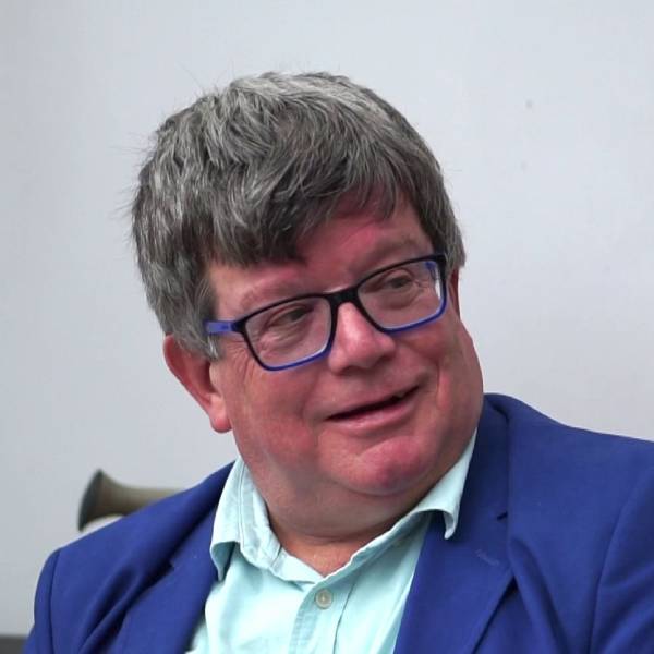 A middle aged white man with dark grey short hair, Antony, wearing a blue suit and blue framed glasses.