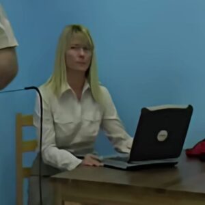 A white, blonde woman wearing a white blouse is sitting at a desk in front of a laptop