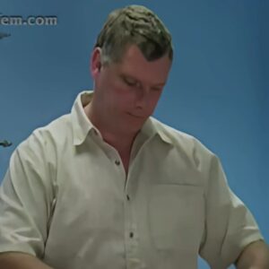 A middle aged white man with dark grey hair, wearing a light coloured shirt and looking down at something off-screen
