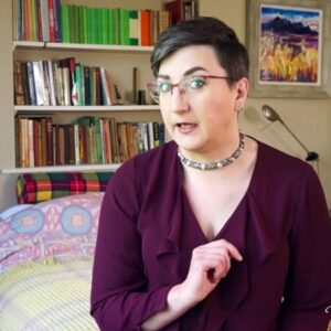 Pandora Blake is wearing a burgandy low cut top and necklace. They are sitting on the bed, looking disapprovingly at the camera