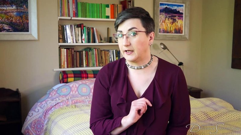 Pandora Blake is wearing a burgandy low cut top and necklace. They are sitting on the bed, looking disapprovingly at the camera