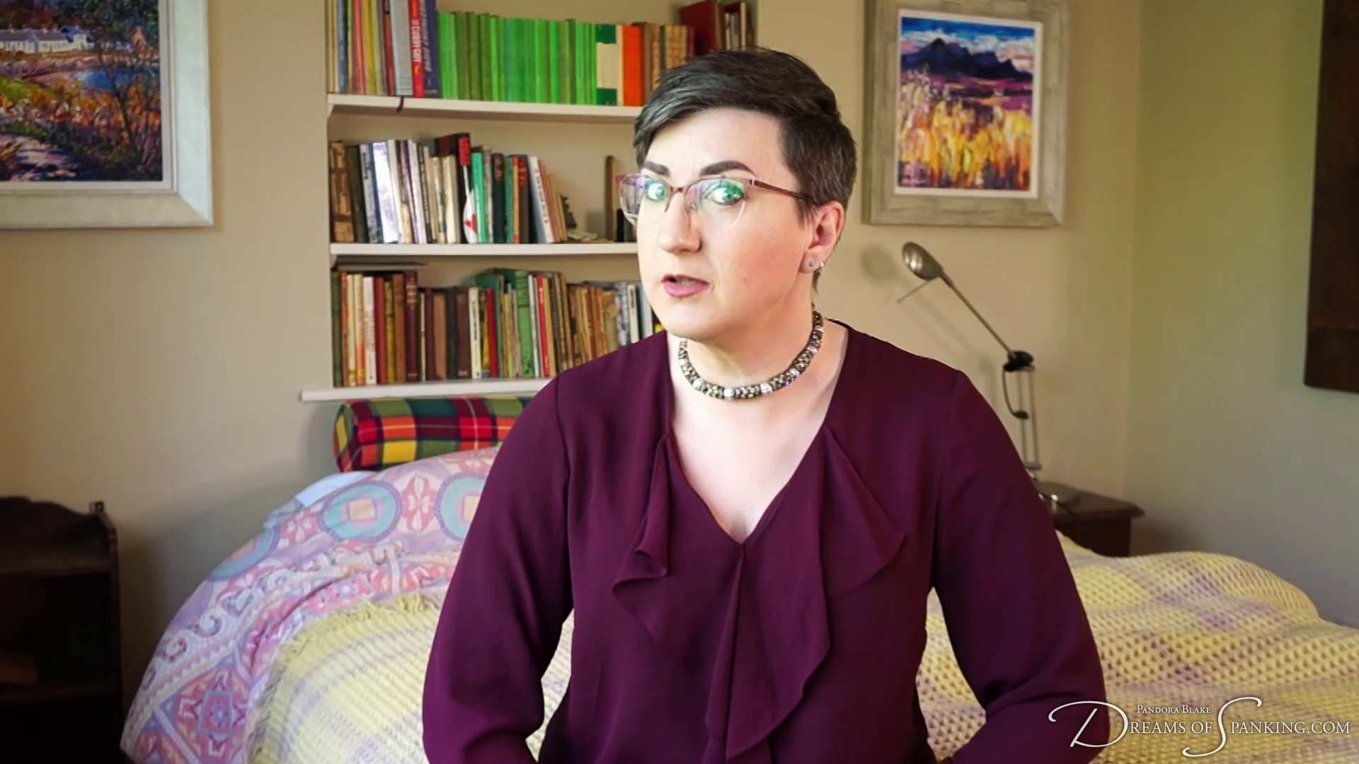 Pandora Blake is wearing a burgandy low cut top and necklace. They are sitting on the bed, looking disapprovingly at the camera