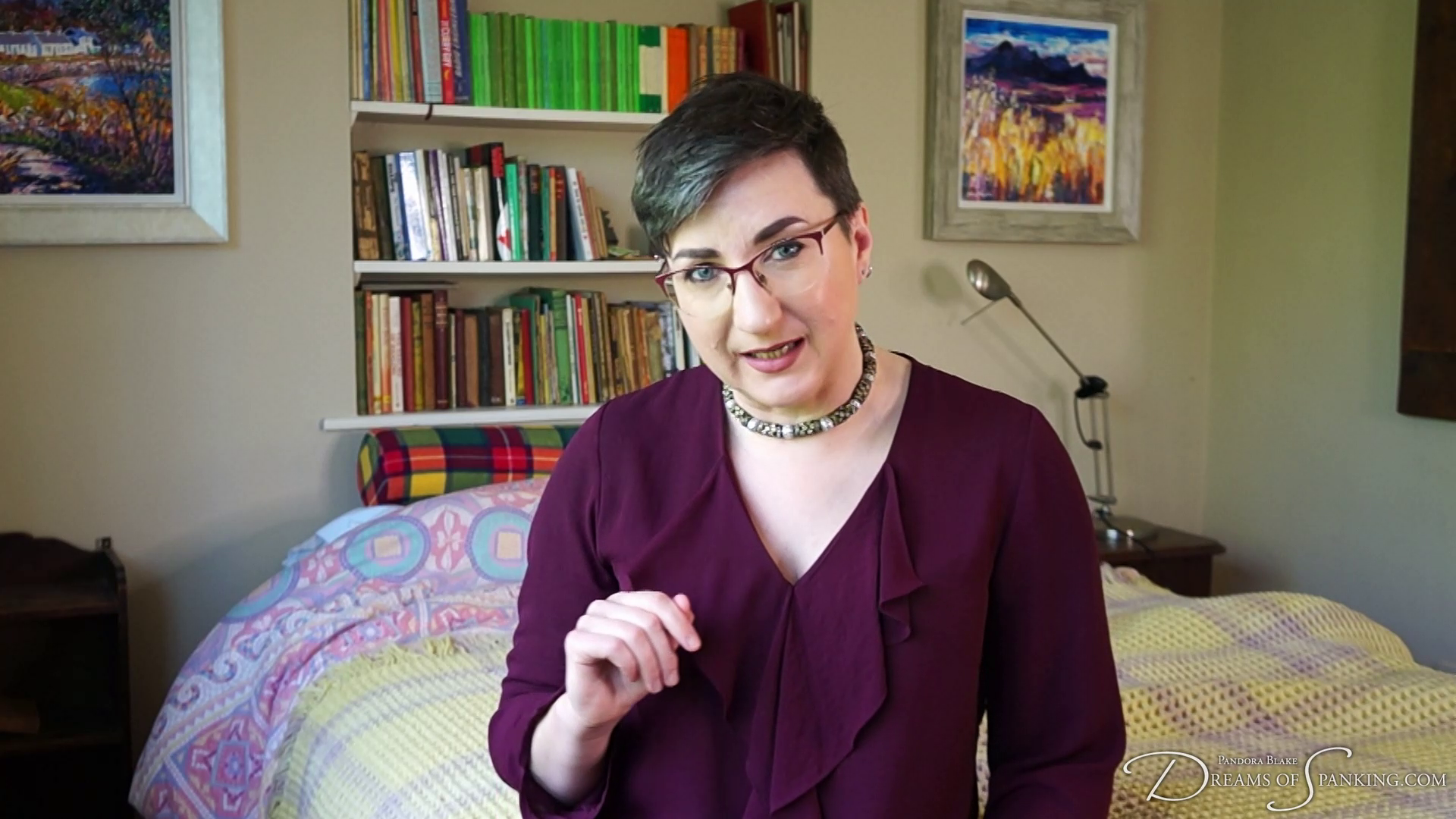 Pandora Blake is wearing a burgandy low cut top and necklace. They are sitting on the bed, looking disapprovingly at the camera